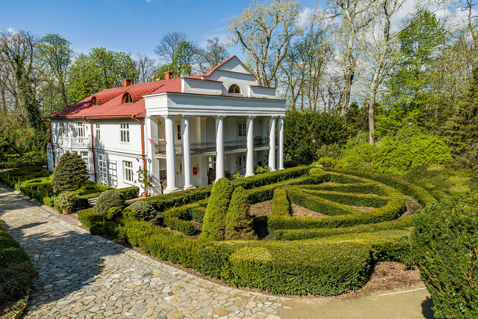 Główny budynek Arboretum, z przodu kolumny. Budynek koloru białego z dachem koloru czerwonawego. Z przodu budynku ułożone w półkręgu żywopłoty.